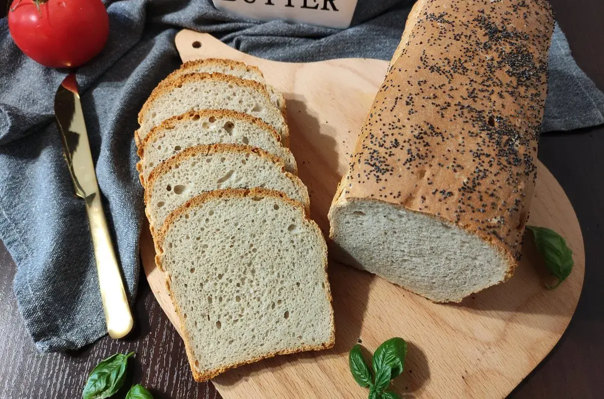 Chleb pszenno żytni z makiem, pokrojony w kromki, leży obok całego bochenka na desce. Obok pomidor, nóż i listki bazylii.