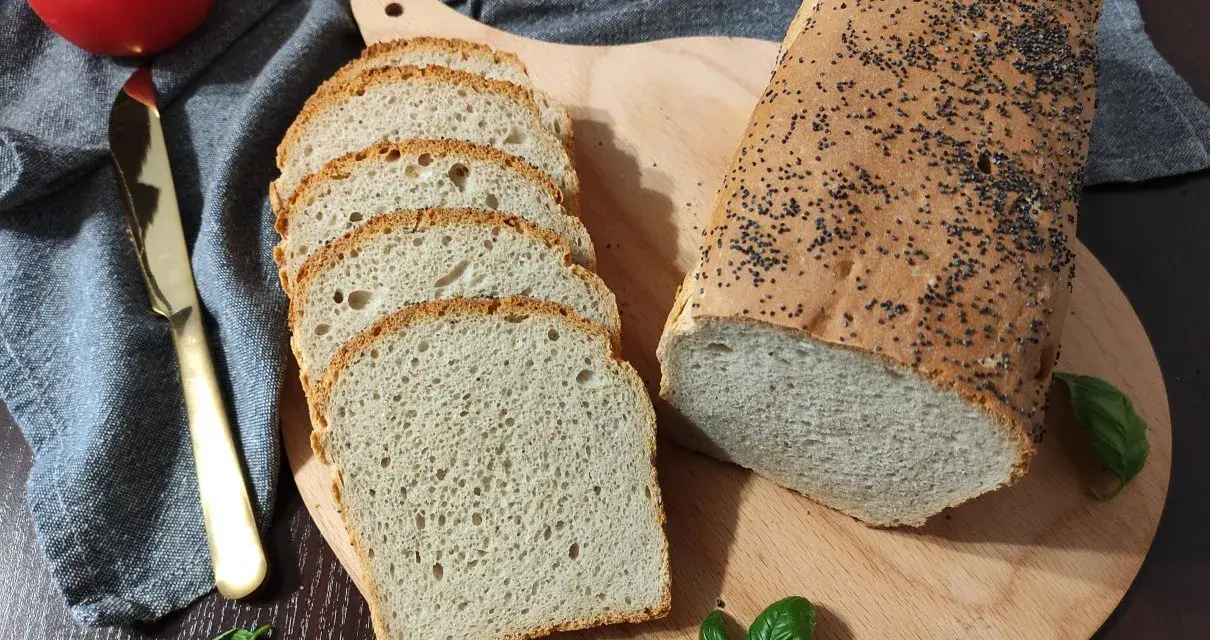 Chleb pszenno żytni z makiem, pokrojony w kromki, leży obok całego bochenka na desce. Obok pomidor, nóż i listki bazylii.