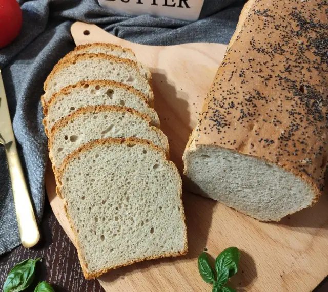 Chleb pszenno żytni z makiem, pokrojony w kromki, leży obok całego bochenka na desce. Obok pomidor, nóż i listki bazylii.