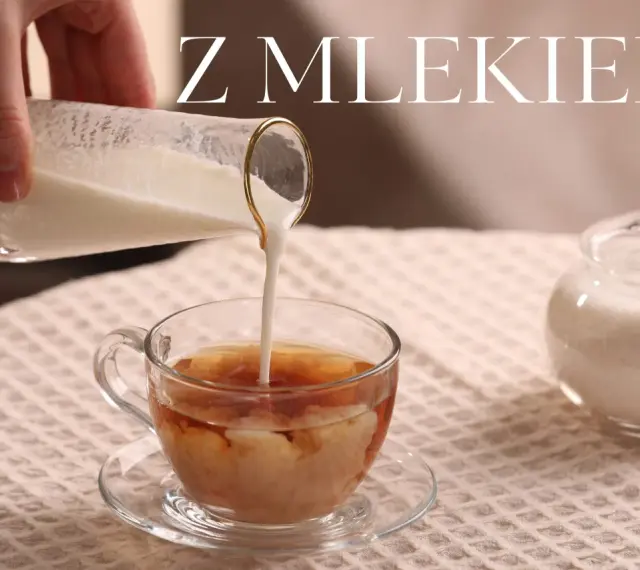Ręka wylewa mleko do szklanki z herbatą, tworząc kremowe wzory. Obok stoi cukiernica. Idealna herbata z mlekiem na chłodny dzień.