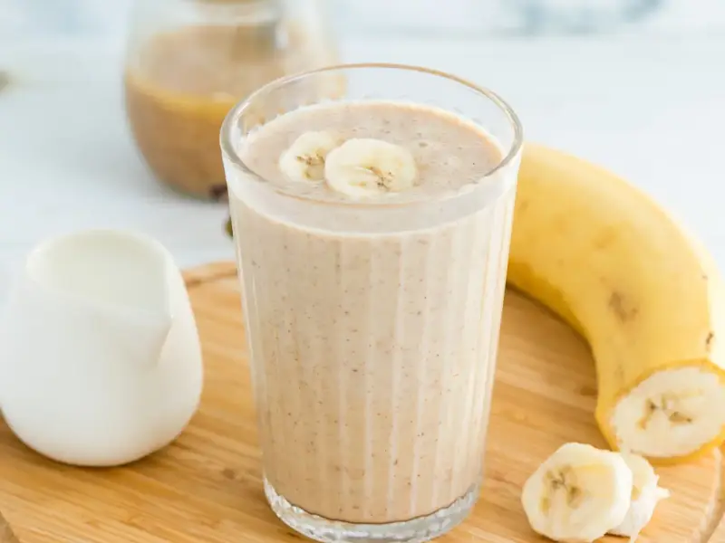Pyszny koktajl bananowy, udekorowany plasterkami banana, obok dojrzałego banana i dzbanka z mlekiem. Idealny na orzeźwiający początek dnia.