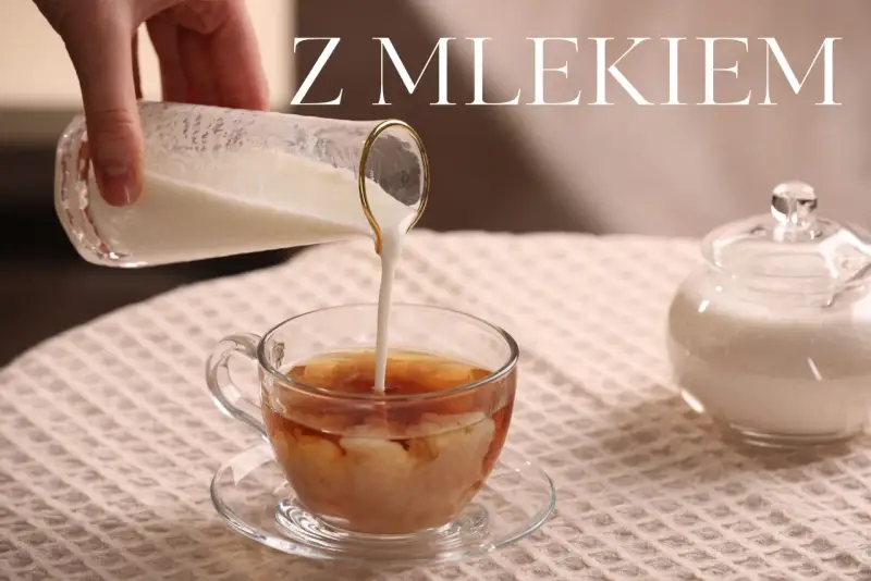Ręka wylewa mleko do szklanki z herbatą, tworząc kremowe wzory. Obok stoi cukiernica. Idealna herbata z mlekiem na chłodny dzień.